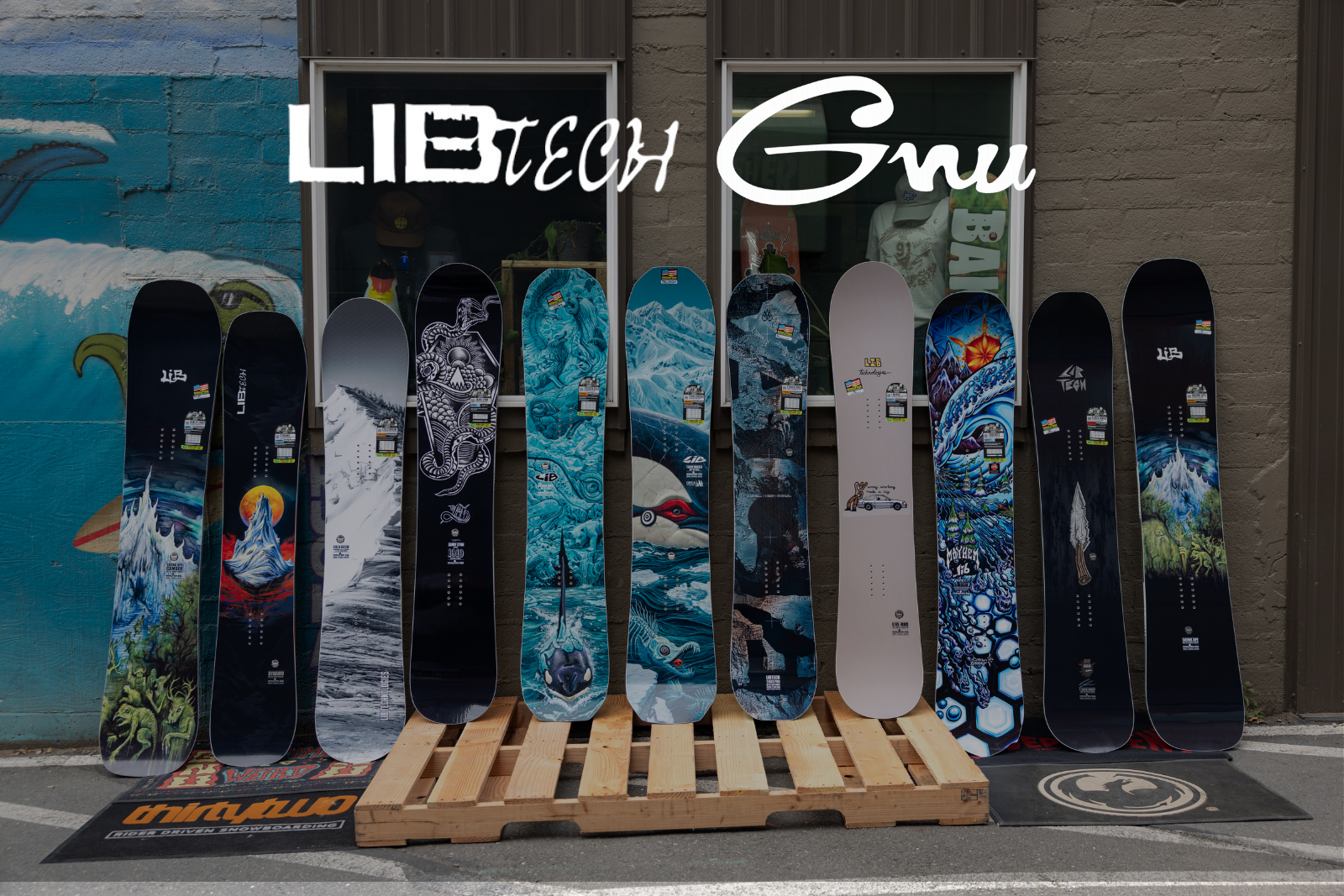 A lineup of ten snowboards with various graphic designs displayed upright on a wooden pallet in front of a brick wall storefront. The brand names 'LIBTECH' and 'Gnu' are prominently displayed in white text at the top.