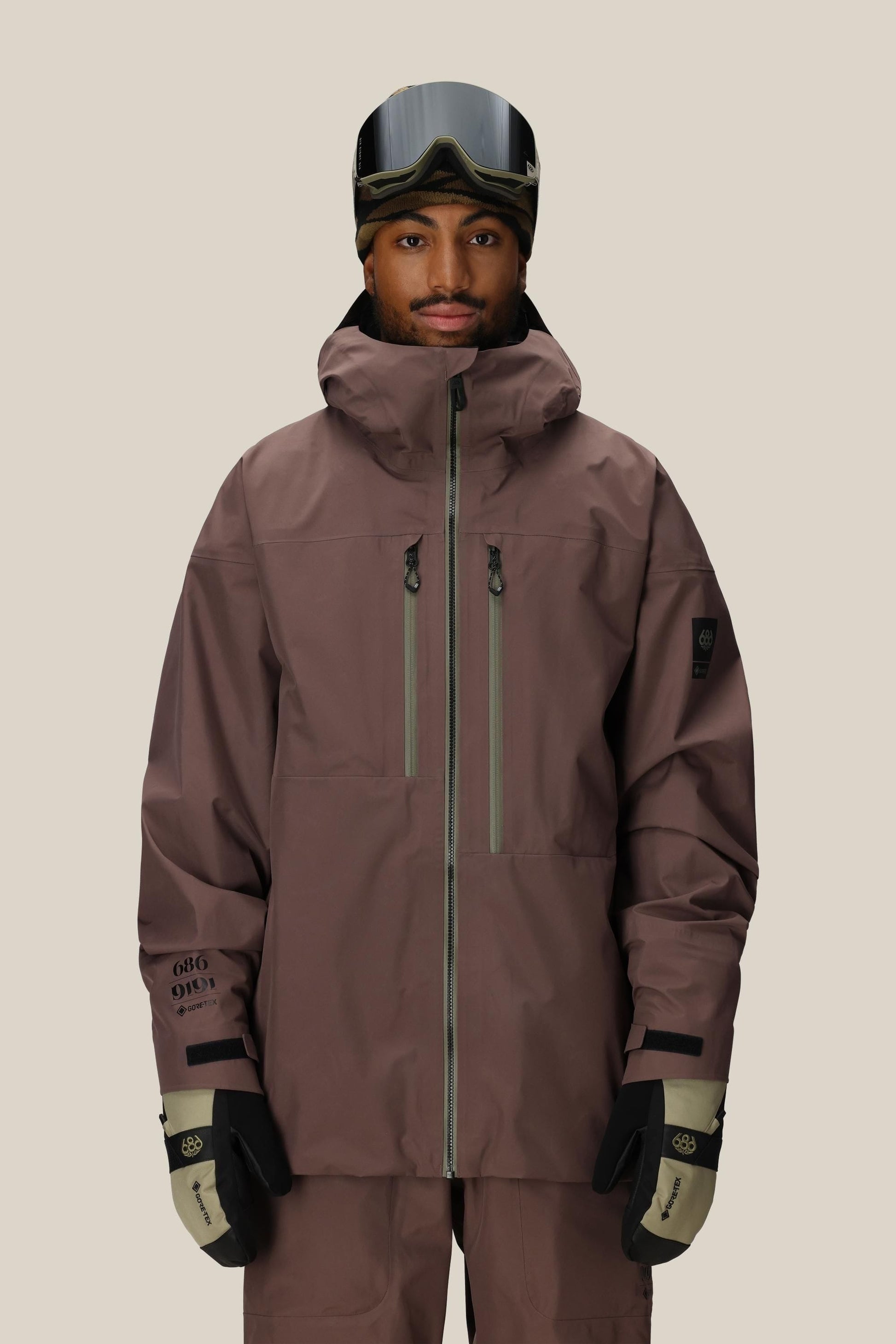 Person wearing a 686 GORE-TEX 3L ATV Thermagraph® Jacket 1 snow jacket, pants, and goggles on a beige background.