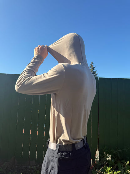 A person outdoors wearing a WWS Baselayer long-sleeve hooded top in the stone colorway, pulling the hood up over the head. The photo is taken in bright sunlight against a clear blue sky and a dark green fence.